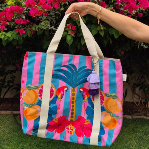 A hand holding a beach bag in the garden in front of pink flowers. The Beach Bag - Frangipani Parrots by Natural Selection is a colourful tote crafted from poly canvas, showcasing beige handles, a vibrant palm tree design with parrots, oranges, and pink hibiscus on pink and teal stripes. It features waterproof lining and a tassel charm on one handle.