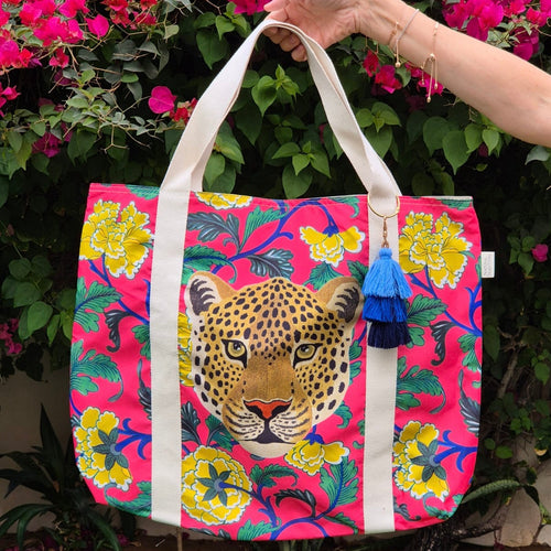 Lady holding the Natural Selection's Beach Bag - Boho Taj by the handles in a garden. The bag showcases a vibrant pink background with yellow flowers and green leaves, all featuring an intricately detailed leopard face. This bold, colourful design is perfect for the beach or for a pool day.