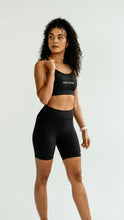 Load image into Gallery viewer, Woman wearing a black sports bra and high-waisted black Koko Actove shorty shorts with branding on a white background.
