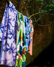Load image into Gallery viewer, The Bobums Beach Towel - Pink &amp; Black Palms, showcasing tropical prints with palm trees and abstract patterns in vibrant blue, green, orange, and pink hues, hangs drying against a rock backdrop.
