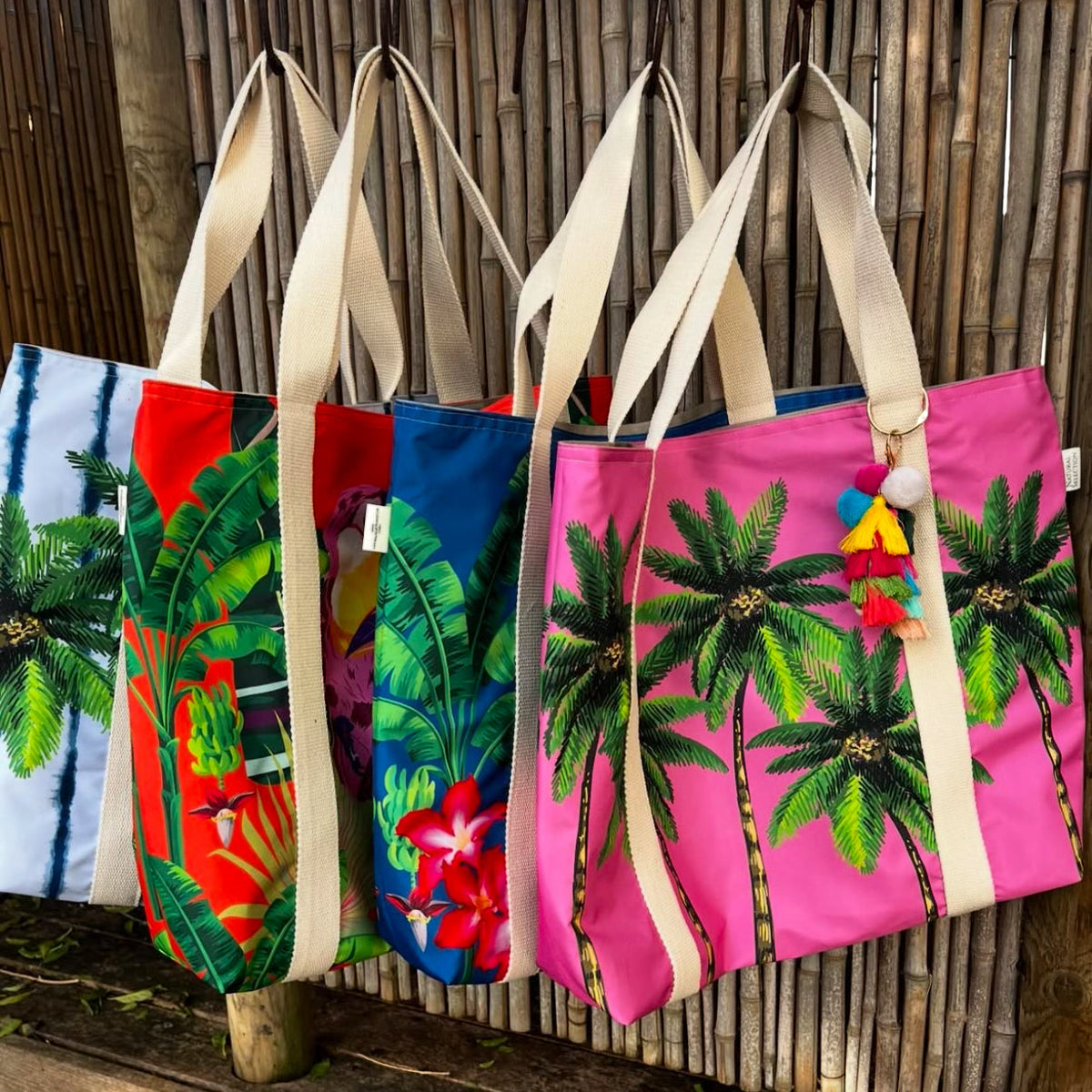 Four Natural Selection beach bags hanging  - colourful totes crafted from poly canvas, showcasing beige handles and a vibrant design . They feature waterproof lining and a tassel charm on one handle.