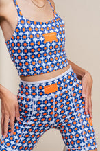 Load image into Gallery viewer, A person wears a vibrant Koko Active set featuring &quot;Shorty Shorts - The Court&quot; with a blue and orange geometric pattern. The tank pairs perfectly with the high-waisted shorts, both having an orange label. The UV50 protection fabric offers thoughtful design against a neutral backdrop.
