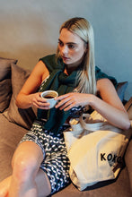 Load image into Gallery viewer, A blonde woman sits on a sofa with a coffee cup, wearing an ethically designed black-and-white patterned outfit and a green sweater draped over her shoulders. On her lap is a beige tote bag labeled &quot;Koko Active.
