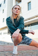 Load image into Gallery viewer, Kneeling outdoors, a confident woman with long blonde hair and a green sweatshirt wears Koko Active&#39;s Shorty Shorts - The Retro Grade. Posed in front of a modern building, her style embodies chic, ethically designed fashion.
