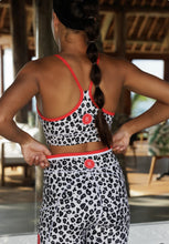 Load image into Gallery viewer, A person with a long braid stands indoors, sporting Koko Active&#39;s Extra Shorty Shorts - Step Up in a leopard-print with red trim. They face a large mirror, hands on hips.
