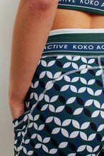 Load image into Gallery viewer, A close-up shows someone wearing Koko Active&#39;s Shorty Shorts in Retro Grade print, featuring interlocking circles in blue, black, and white. The ethically designed geometric pattern has &quot;KOKO ACTIVE&quot; on the waistband, with the person’s hand resting inside a pocket.
