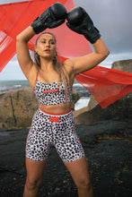 Load image into Gallery viewer, A person in leopard print Extra Shorty Shorts - Step Up by Koko Active poses with boxing gloves raised, standing on rocky terrain near the ocean. A red translucent fabric waves in the background, while UV50 protection gear ensures they&#39;re ready for any weather under an overcast sky.
