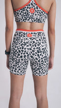 Load image into Gallery viewer, A person models Koko Active&#39;s ethically produced leopard print sports bra and Extra Shorty Shorts - Step Up, featuring a red waistband, back branding for UV50 protection. The individual stands turned away, highlighting a black smartwatch against a plain light background.
