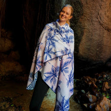Load image into Gallery viewer, A smiling person wrapped in a Bobums Beach Towel - Pink &amp; Black Palms stands by a rocky wall, sunlight highlighting their face naturally.
