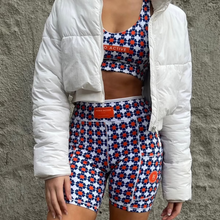 Load image into Gallery viewer, A person wears Koko Active&#39;s chic white puffer jacket over a matching sports bra and The Court shorty shorts. The ensemble, adorned with orange and blue flowers on a crisp white background, showcases UV50 protection fabric for style and functionality.
